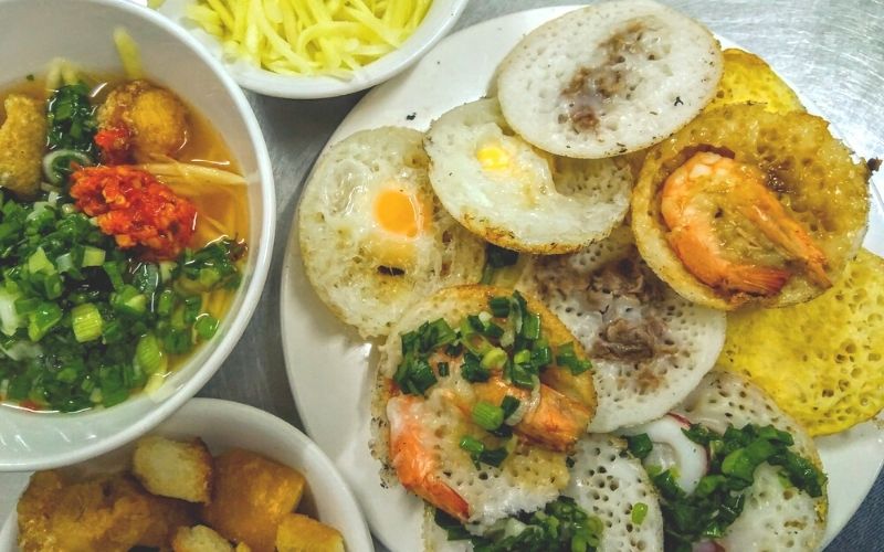 Đĩa bánh căn trứng, tôm, mực bên các chén nước chấm và xoài